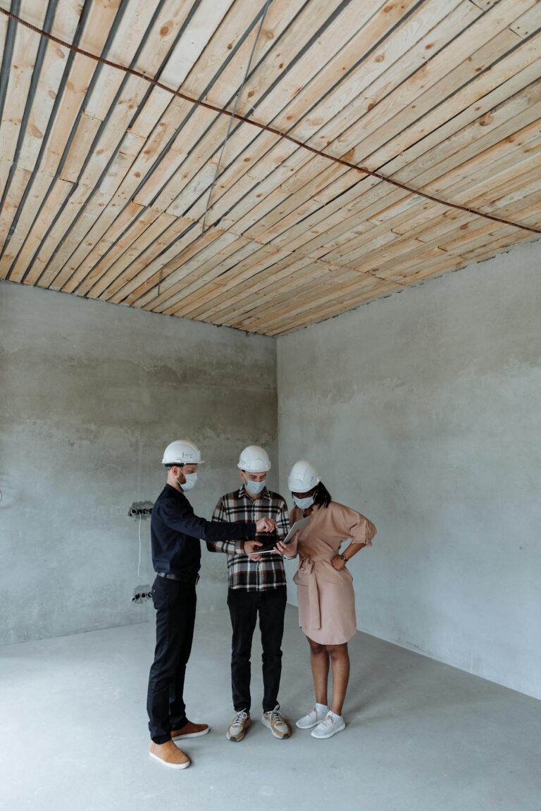 Engineers wearing hard hats review building plans in an unfinished room, discussing construction strategy.