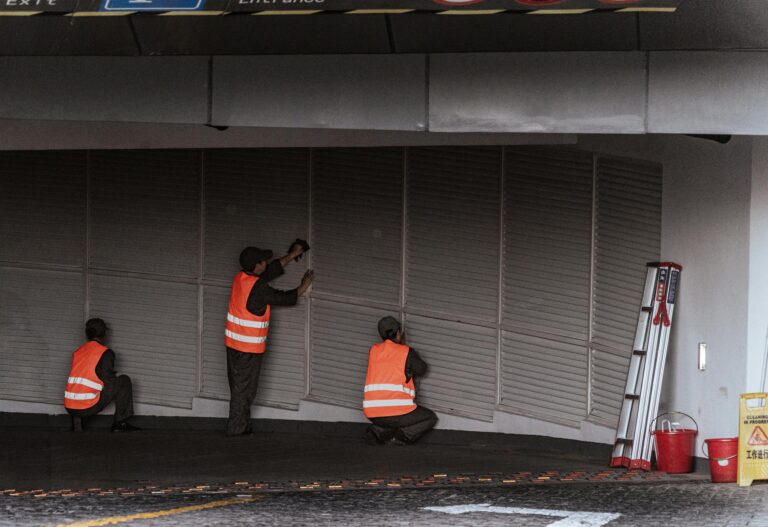 Three workers in safety vests install security shutters at an indoor entrance.